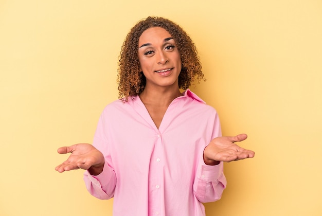 Young latin transsexual woman isolated on yellow background makes scale with arms, feels happy and confident.