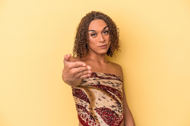 Молодая латинская транссексуальная женщина изолирована на желтом фоне, протягивая руку перед камерой в приветственном жесте.