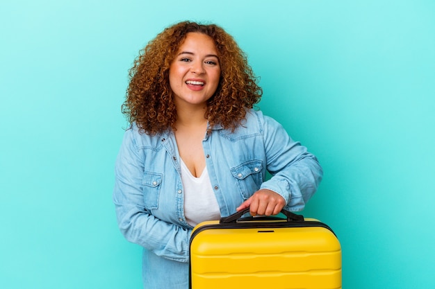 Young latin traveler curvy woman holding a suitcase isolated on blue background laughing and having fun.