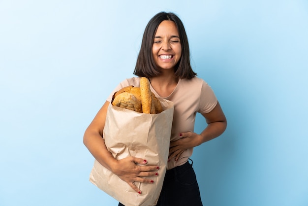 Young latin woman buying some breads isolated on blue smiling a lot