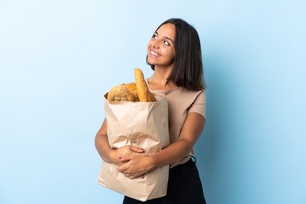 Молодая латинская женщина покупает хлеб, изолированную на синем, думая об идее, глядя вверх