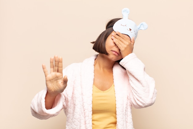 Young latin woman covering face with hand and putting other hand up front to stop camera, refusing photos or pictures