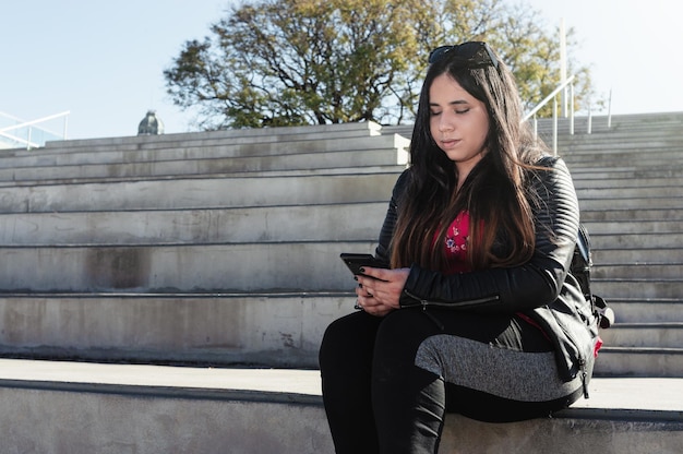 Young latin woman dressed in black outdoors sitting on a grand staircase using her cell phone