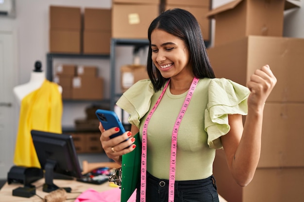 Young latin woman dressmaker with winner expression using smartphone at designer studio