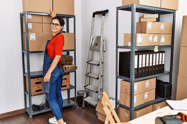 Young latin woman ecommerce business worker organizing packages on shelving at office