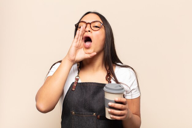 Young latin woman feeling happy, excited and positive, giving a big shout out with hands next to mouth, calling out