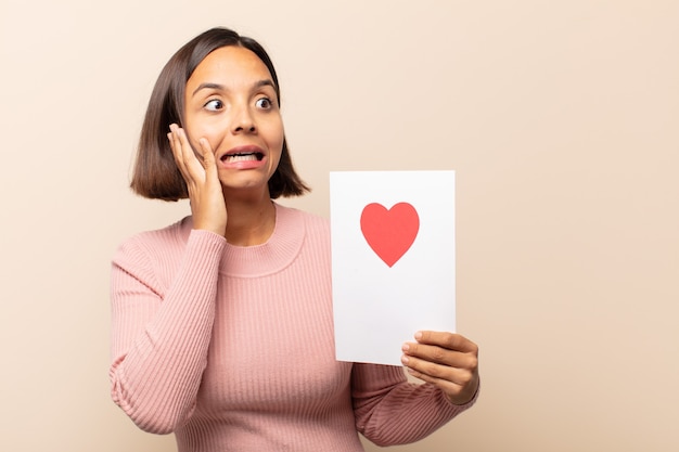 Giovane donna latina che si sente felice, eccitata e sorpresa, guardando di lato con entrambe le mani sul viso