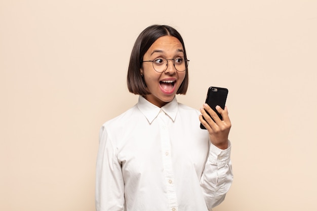 Young latin woman feeling happy, excited and surprised, looking to the side with both hands on face