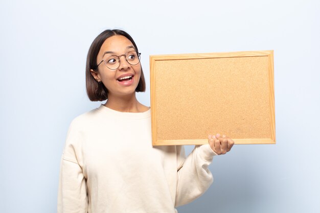 Young latin woman feeling happy, excited and surprised, looking to the side with both hands on face