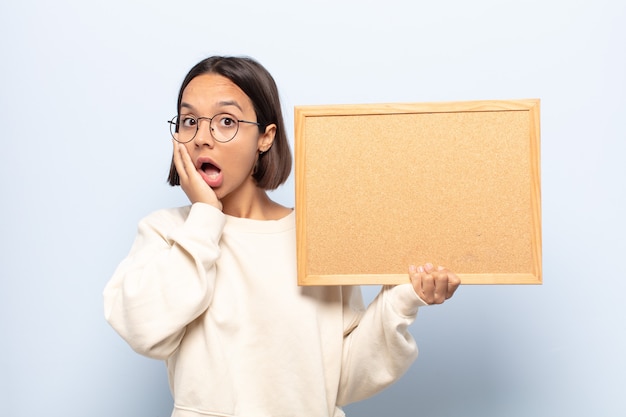 Young latin woman feeling shocked and scared, looking terrified with open mouth and hands on cheeks