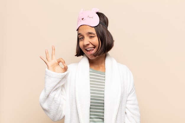 Young latin woman feeling successful and satisfied, smiling with mouth wide open, making okay sign with hand