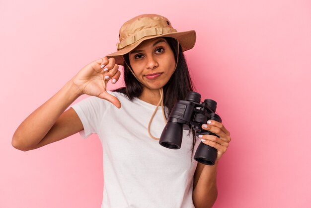 Young latin woman holding binoculars isolated on pink background showing a dislike gesture, thumbs down. Disagreement concept.