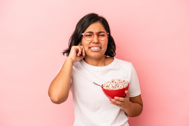 Young latin woman holding cereals isolated on pink background covering ears with hands.