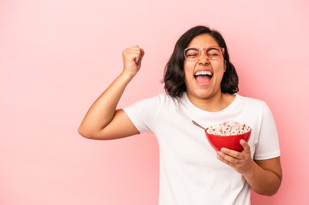 Young latin woman holding cereals isolated on pink background raising fist after a victory, winner concept.