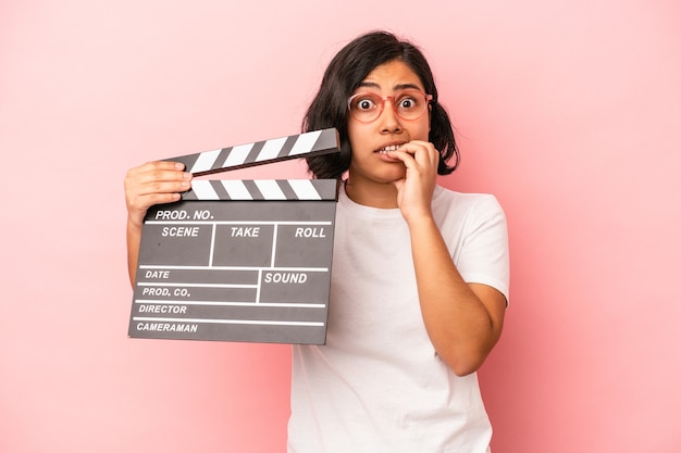Young latin woman holding clapperboard isolated on pink background biting fingernails, nervous and very anxious.