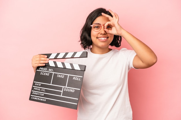 Young latin woman holding clapperboard isolated on pink background excited keeping ok gesture on eye.