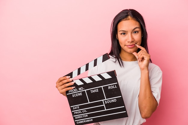 Young latin woman holding clapperboard isolated on pink background  relaxed thinking about something looking at a copy space.