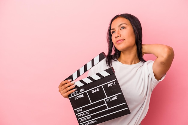 Young latin woman holding clapperboard isolated on pink background  touching back of head thinking and making a choice