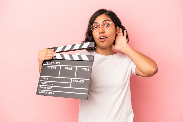 Young latin woman holding clapperboard isolated on pink background trying to listening a gossip.