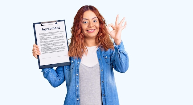 Young latin woman holding clipboard with agreement document doing ok sign with fingers, smiling friendly gesturing excellent symbol