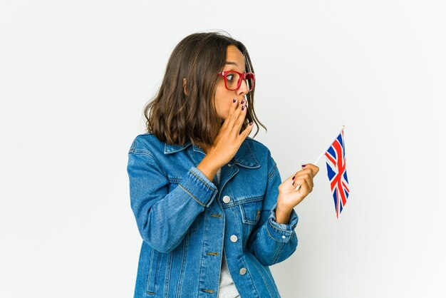 Young latin woman holding a english flag isolated on white wall is saying a secret hot braking news and looking aside