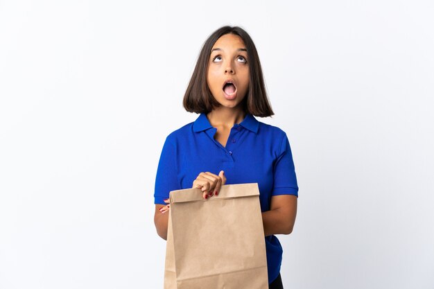 Молодая латинская женщина, держащая продуктовую сумку для покупок изолирована на белом, глядя вверх и с удивленным выражением лица