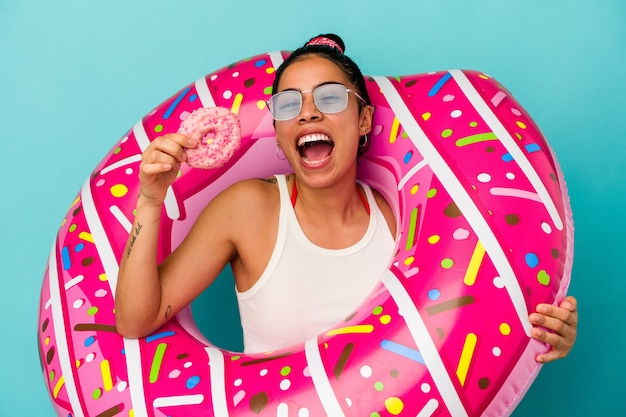 Young latin woman holding an inflatable donut eating a donut isolated on blue background