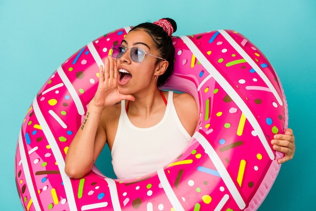 Young latin woman holding an inflatable donut isolated on blue background shouting and holding palm near opened mouth.