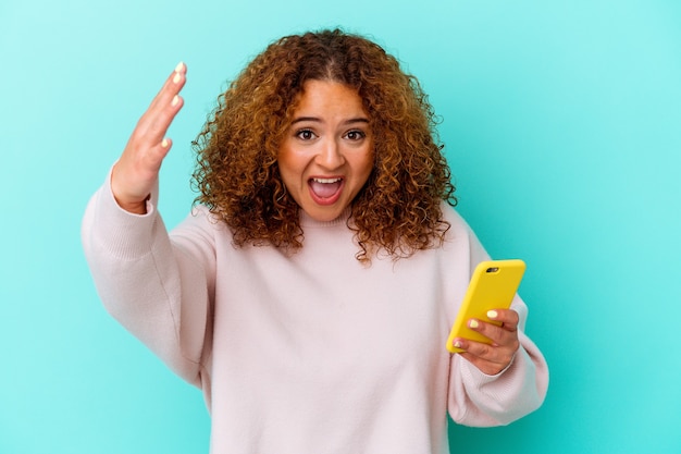 Молодая латинская женщина, держащая мобильный телефон, изолирована на синем фоне, получая приятный сюрприз, возбужденный и поднимающий руки.