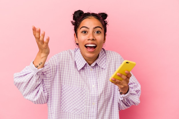 Young latin woman holding a mobile phone isolated on pink background receiving a pleasant surprise, excited and raising hands.