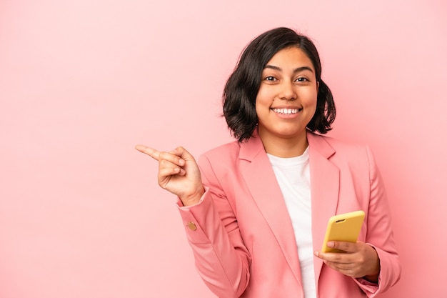 Young latin woman holding mobile phone isolated on pink background smiling and pointing aside, showing something at blank space.