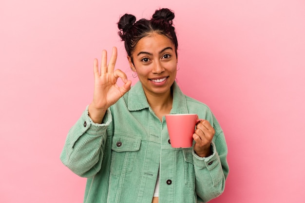 ピンクの背景に分離されたマグカップを持っている若いラテン女性は陽気で自信を持ってOKジェスチャーを示しています。