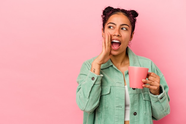 Young latin woman holding a mug isolated on pink background is saying a secret hot braking news and looking aside