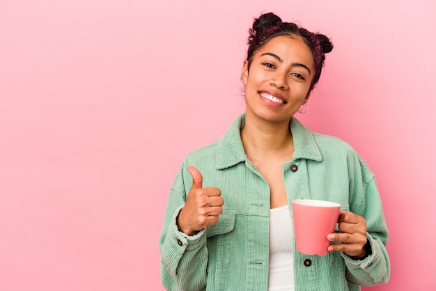 Young latin woman holding a mug isolated on pink background smiling and raising thumb up