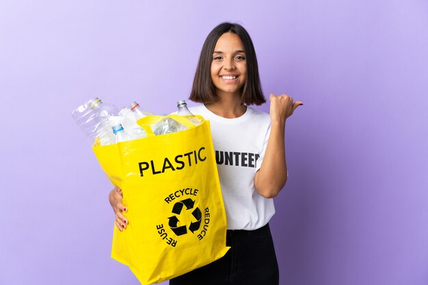 Young latin woman holding a recycling bag full of paper to recycle isolated on purple pointing to the side to present a product