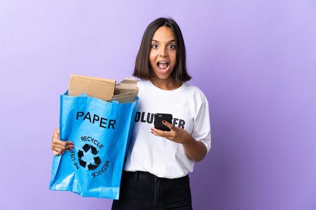 驚いて、メッセージを送信する紫に分離されたリサイクルに紙の完全なリサイクルバッグを保持している若いラテン女性