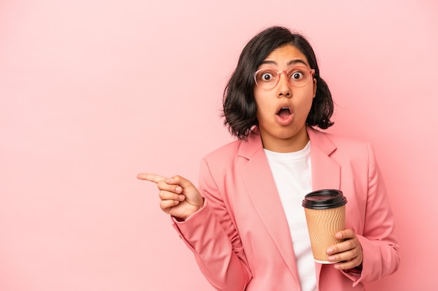 Young latin woman holding take away coffee isolated on pink background pointing to the side