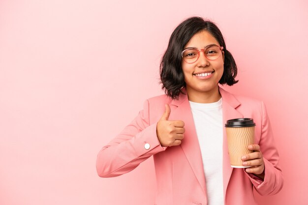 保持している若いラテン女性は笑顔と親指を上げてピンクの背景に分離されたコーヒーをテイクアウト