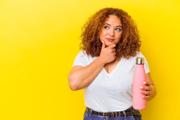 Young latin woman holding a thermos isolated on yellow background looking sideways with doubtful and skeptical expression.