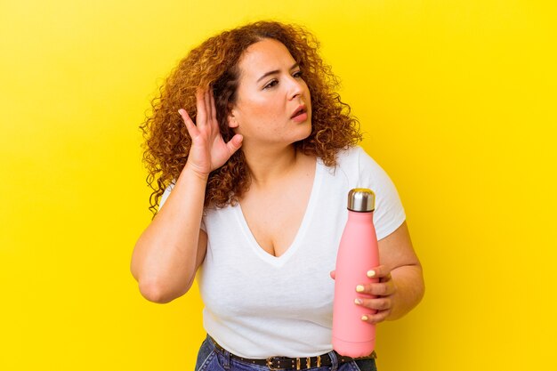 Young latin woman holding a thermos isolated on yellow background trying to listening a gossip.