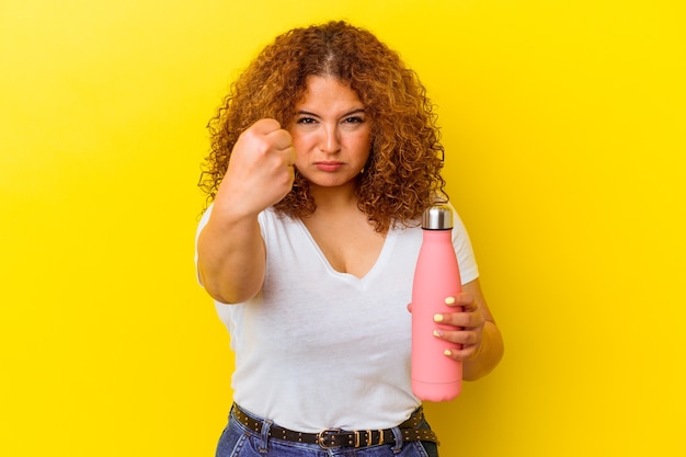 Young latin woman holding a thermos isolated on yellow wall showing fist to front, aggressive facial expression