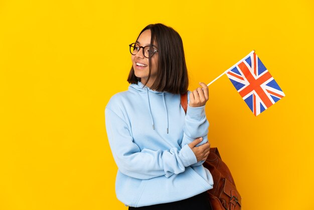 Young latin woman holding an United Kingdom flag isolated on yellow background looking to the side and smiling