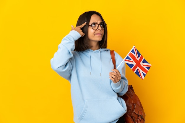 Young latin woman holding an United Kingdom flag isolated on yellow background making the gesture of madness putting finger on the head