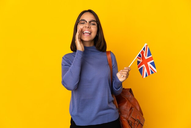 Young latin woman holding an United Kingdom flag isolated on yellow background shouting with mouth wide open