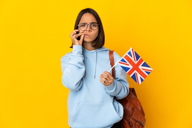 Young latin woman holding an United Kingdom flag isolated on yellow background showing a sign of silence gesture