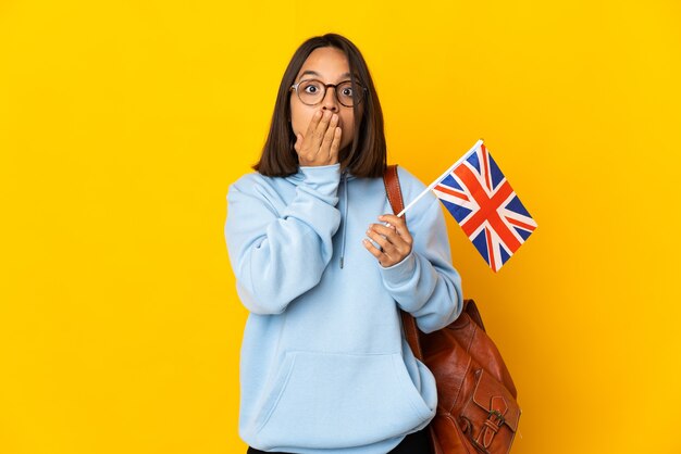 Young latin woman holding an United Kingdom flag isolated on yellow wall covering mouth with hands