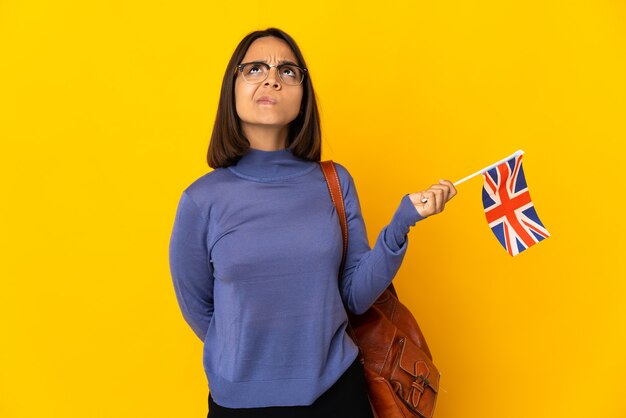 Young latin woman holding an united kingdom flag isolated on yellow wall and looking up