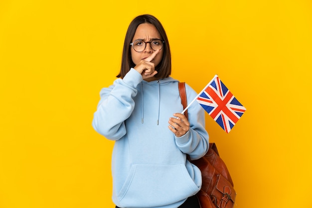 Young latin woman holding an United Kingdom flag isolated on yellow wall showing a sign of silence gesture