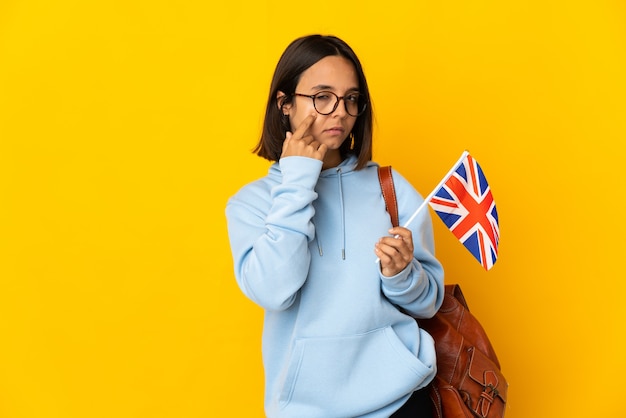 Молодая латинская женщина держит флаг Соединенного Королевства, изолированную на желтой стене, показывая что-то