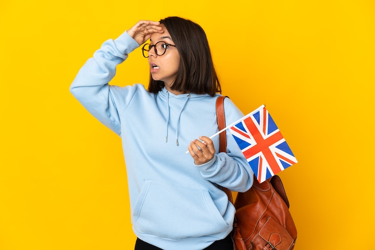 Photo 側面を見ながら驚きの表情で黄色の壁に分離されたイギリスの旗を保持している若いラテン女性
